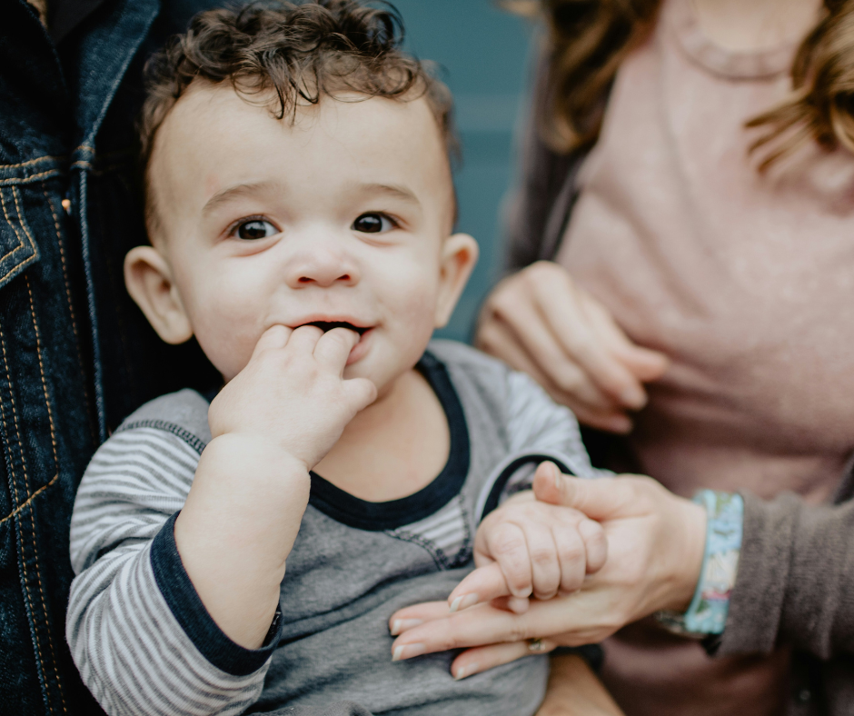 A toddler boy smiles and sucks his fingers as his parents hold him.