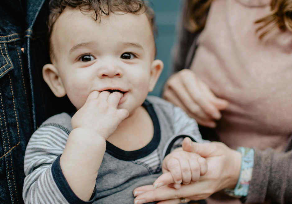 A toddler boy smiles and sucks his fingers as his parents hold him.